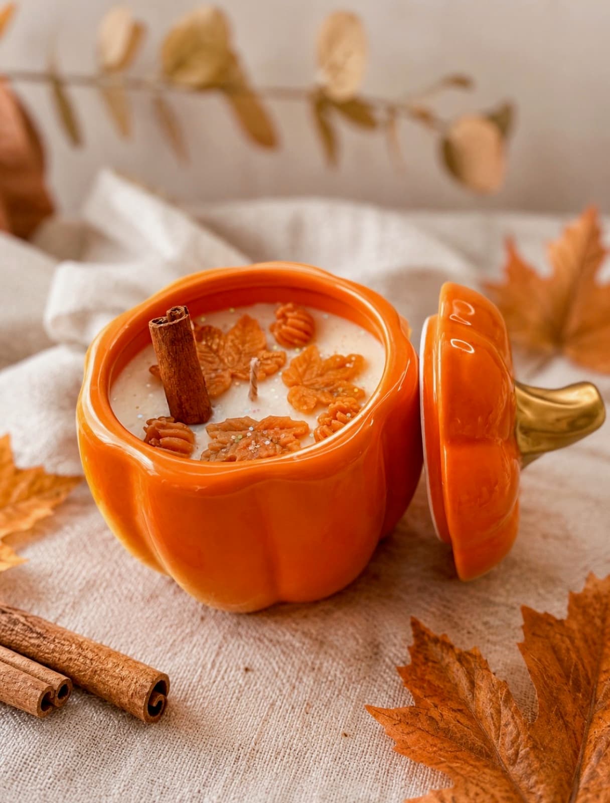Orange ceramic pumpkin candle with a cinnamon stick and wax maple leaves on the surface, lid resting beside it on linen