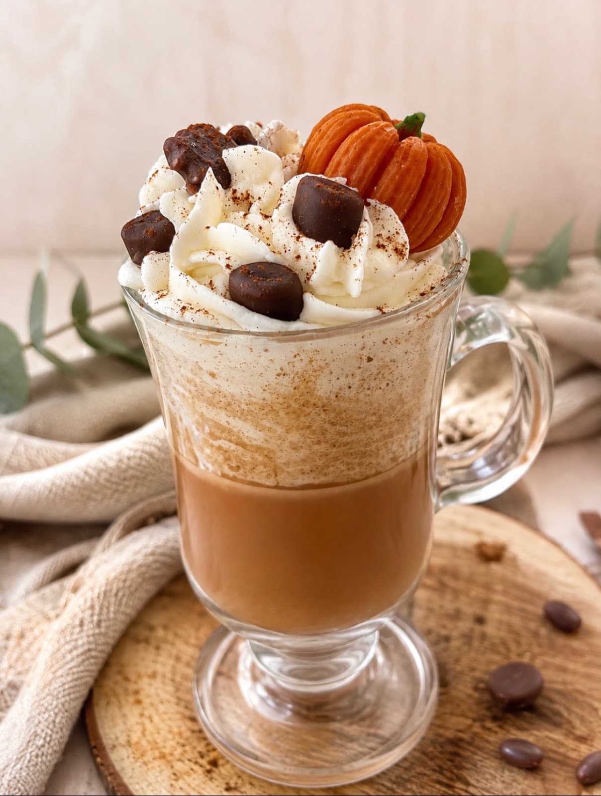 Glass mug candle layered like a latte, topped with piped cream, cocoa dusting, chocolate pieces and a wax pumpkin